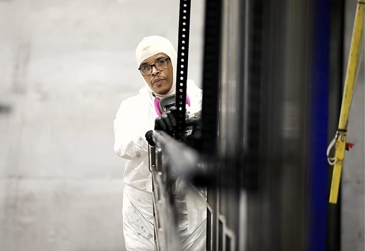 Technician in protective coveralls and respirator preparing a staircase component for powder coat finishing