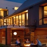 Black Builder Standard Spiral staircase beside a lit fire pit patio at dusk, connecting a deck to the backyard of a modern home