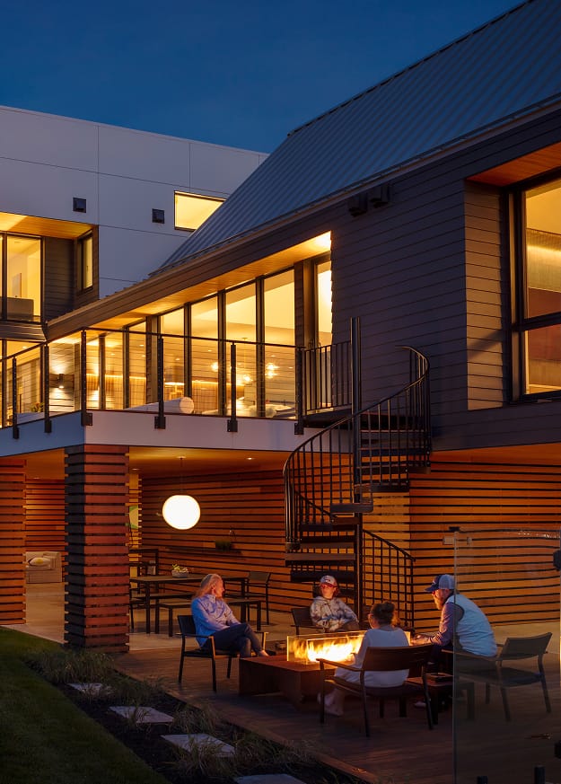 Black Builder Standard Spiral staircase beside a lit fire pit patio at dusk, connecting a deck to the backyard of a modern home