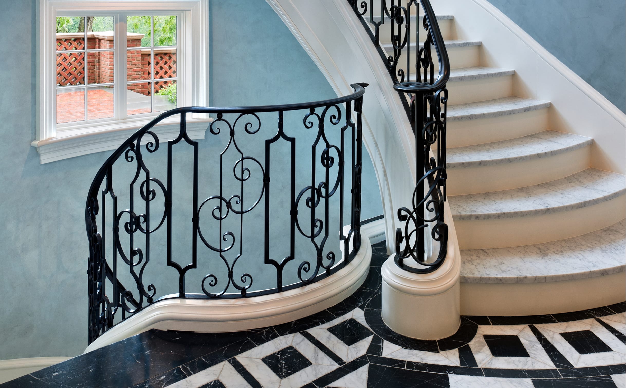 Curved stone staircase with a scrollwork railing in black, next to a window overlooking a brick patio