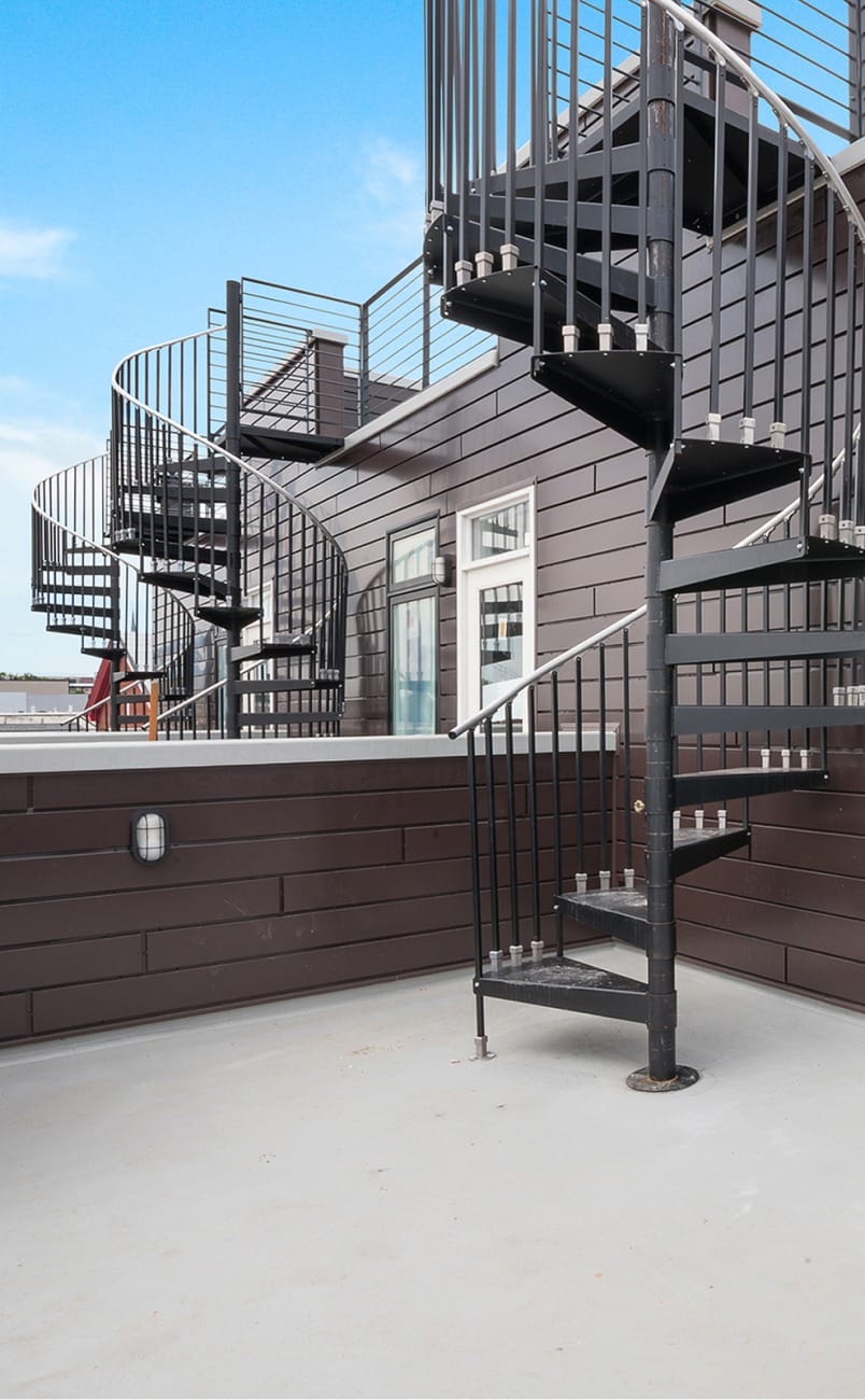 Row of black Builder Standard Spiral series staircases on a rooftop deck of a multi-unit building with dark siding