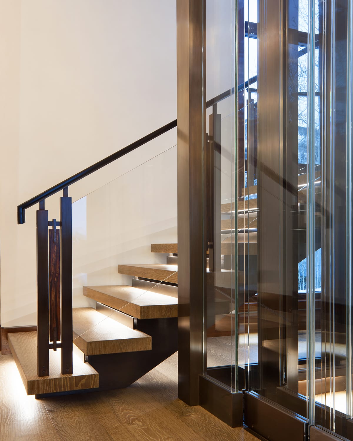 Floating wood staircase with open treads and a dark metal handrail, set against glass elevator panels