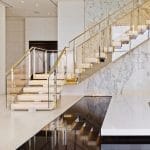 Floating staircase with illuminated onyx treads and a glass and brass railing, mirrored in a dark reflective floor beneath a bookmatched marble wall