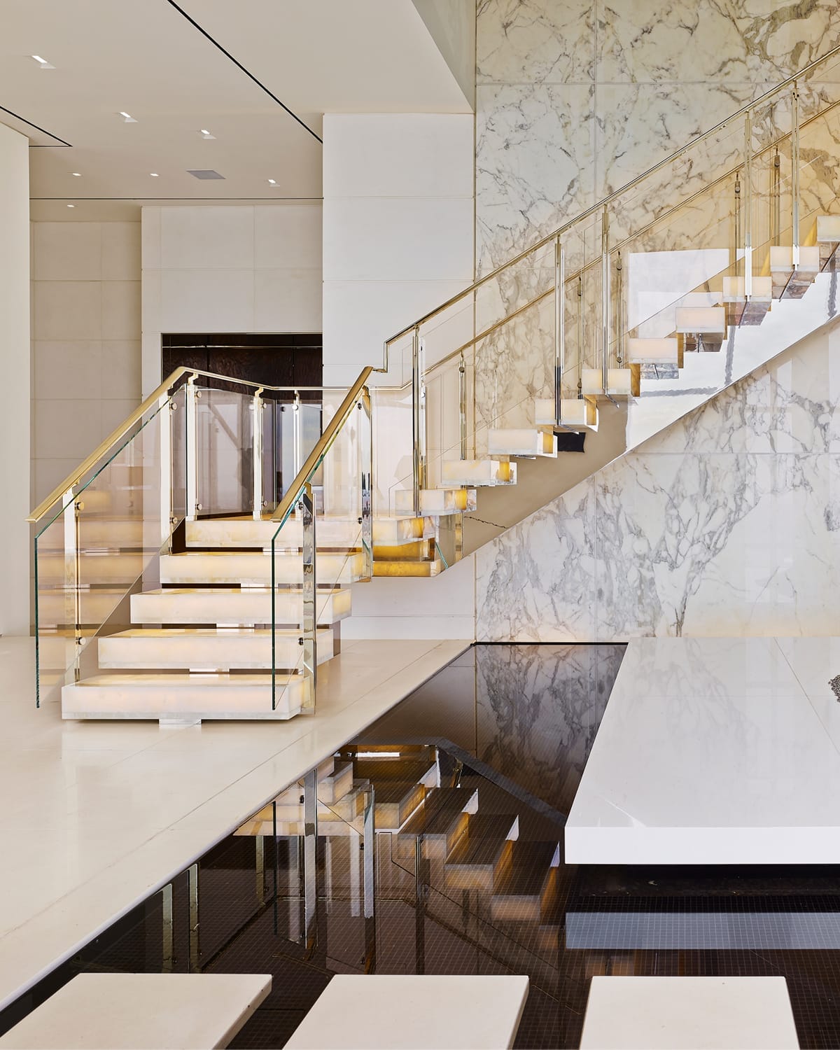 Floating staircase with illuminated onyx treads and a glass and brass railing, mirrored in a dark reflective floor beneath a bookmatched marble wall