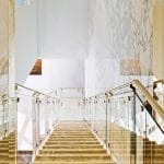 View looking down a staircase with illuminated onyx-clad treads and glass and brass railings, framed by floor-to-ceiling marble walls