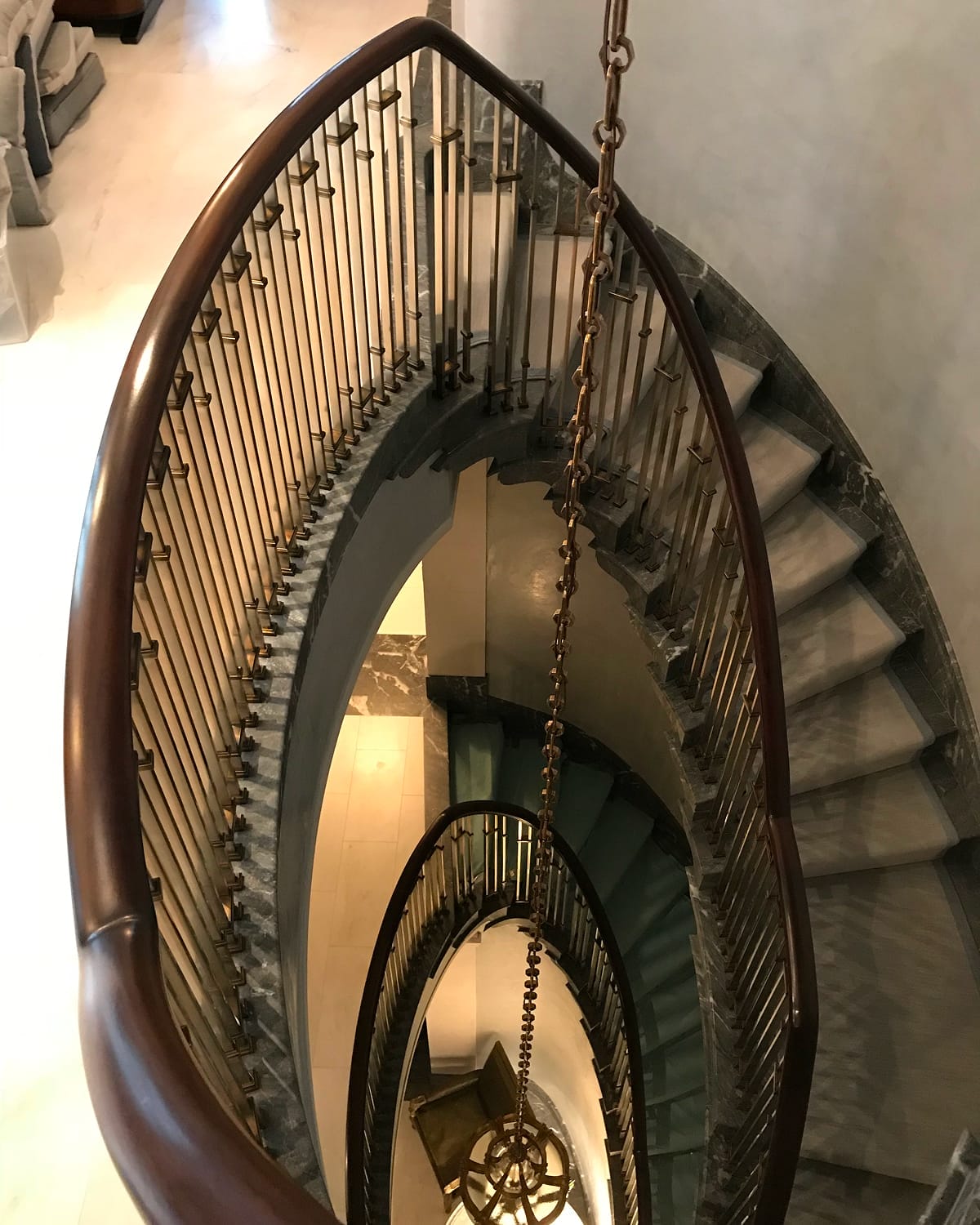 Looking down through an oval stairwell opening, framed by a dark wood handrail and brass-collared spindle railing, with grey marble treads curving around two visible flights and a chandelier chain dropping through the center
