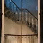 Curved staircase framed through an arched doorway, with a dark wood handrail, brass-collared spindle railing, grey marble treads, and a chandelier chain hanging at center