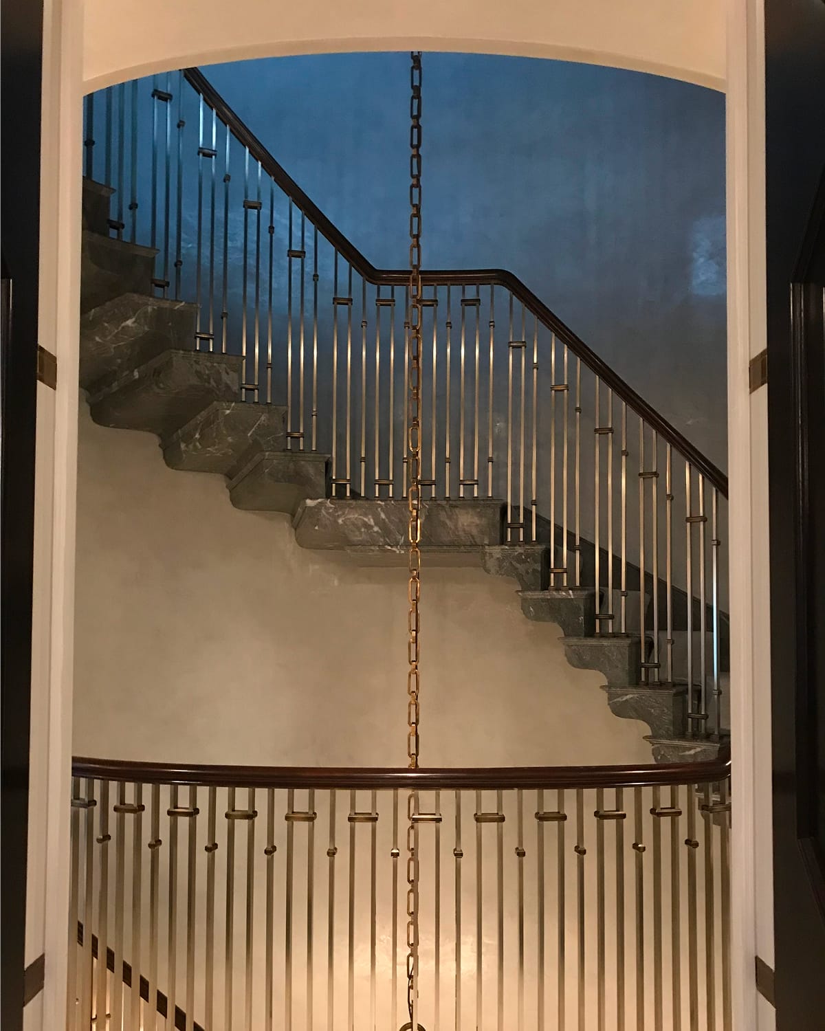 Curved staircase framed through an arched doorway, with a dark wood handrail, brass-collared spindle railing, grey marble treads, and a chandelier chain hanging at center