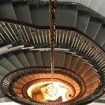 Overhead view down an oval staircase, its grey marble treads and brass-collared spindle railing spiraling around a pendant chandelier glowing at the base