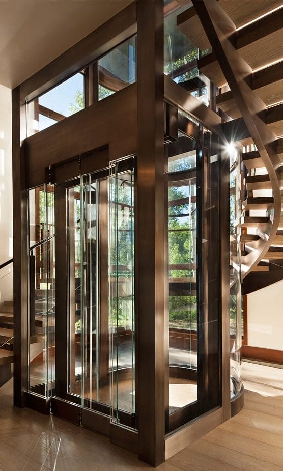 Glass and bronze-framed elevator shaft integrated beside a curved staircase with wood treads