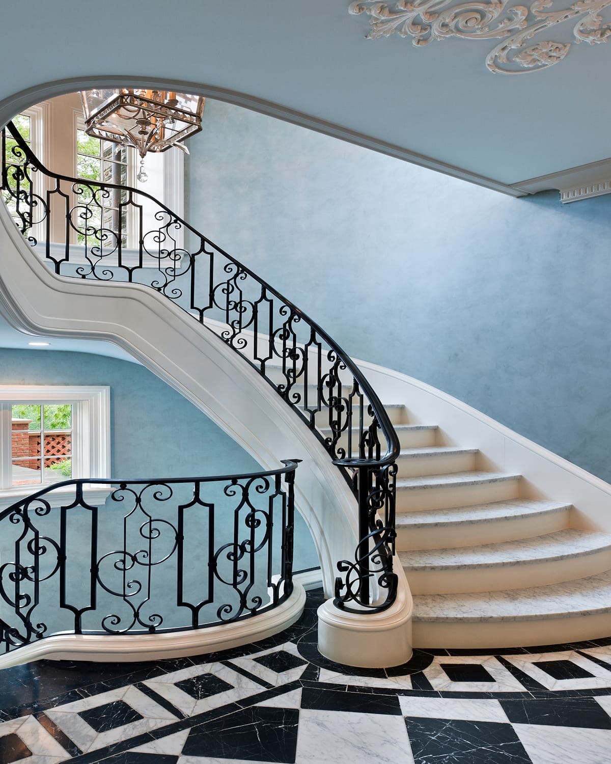 Curved staircase with scrolled iron railing in a Gladwyne, PA entry foyer, with a round wood pedestal table, abstract wall art, and checkered marble flooring