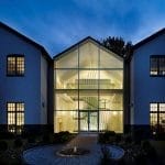 Glass-gabled entrance pavilion lit at night, revealing a sculptural crisscrossed metal installation surrounding a floating staircase inside, framed by a brick courtyard path and fountain