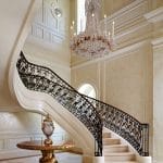 Curved staircase with scrolled iron and gilt-bronze railing descending into a two-story entry hall, lit by a tall crystal chandelier, with a gold-leafed pedestal table at the base