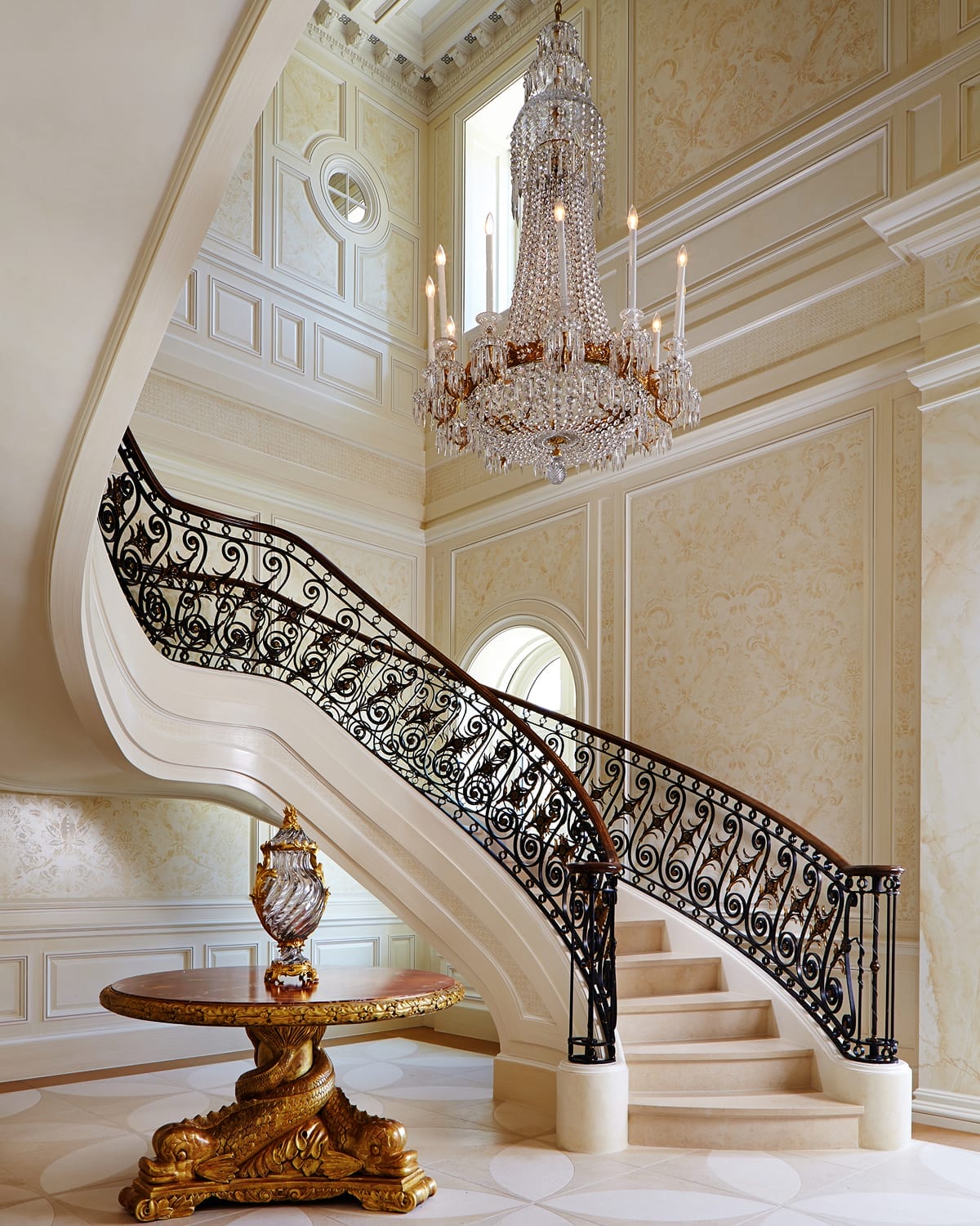 Curved staircase with scrolled iron and gilt-bronze railing descending into a two-story entry hall, lit by a tall crystal chandelier, with a gold-leafed pedestal table at the base