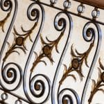 Close detail of a scrolled iron railing panel with gilt-bronze acanthus and rosette accents, set against patterned wallpaper