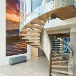 Spiral staircase with light wood treads and a glass and brushed-metal railing, curving past a large sunset seascape photograph and a bench in a bright foyer