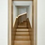 View down a narrow wood-paneled stairwell toward a curved staircase with light wood treads and an illuminated handrail channel