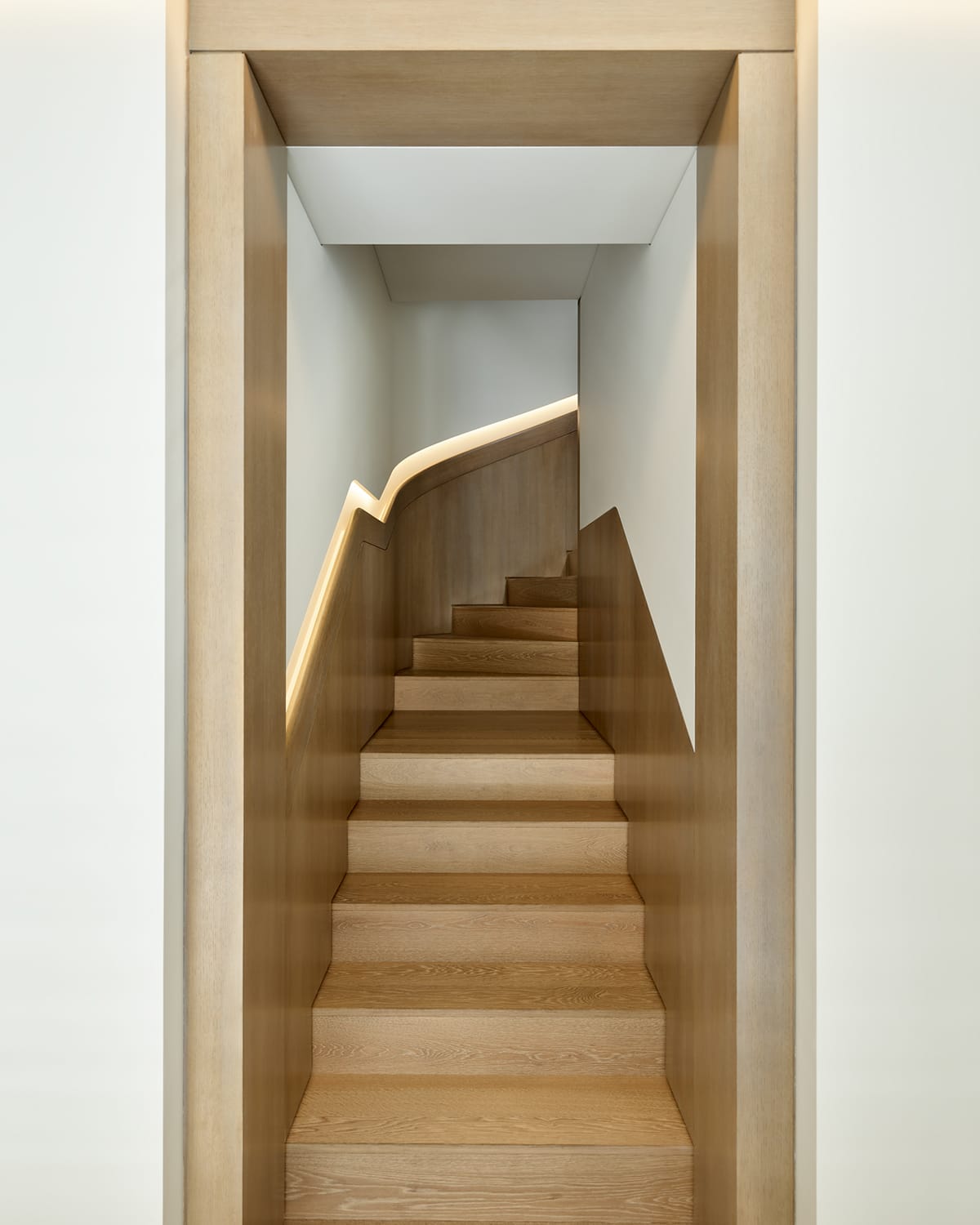 View down a narrow wood-paneled stairwell toward a curved staircase with light wood treads and an illuminated handrail channel