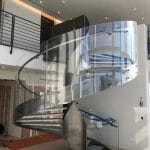 In-progress installation of a spiral staircase, showing the brushed-metal structure and glass railing partially complete, with construction materials and floor protection visible