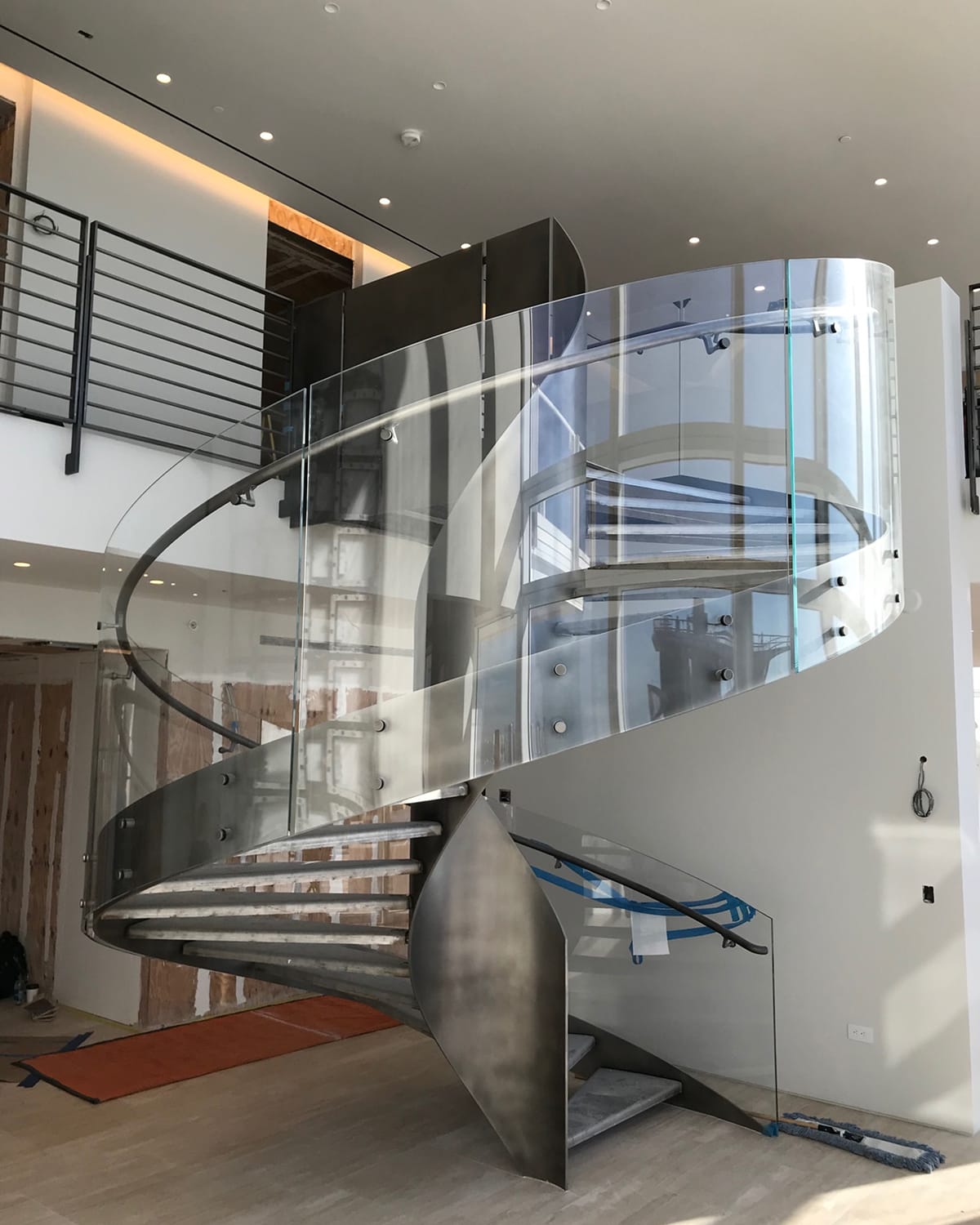 In-progress installation of a spiral staircase, showing the brushed-metal structure and glass railing partially complete, with construction materials and floor protection visible