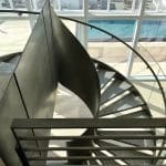 Spiral staircase with wood treads and a glass and brushed-metal railing, beside floor-to-ceiling windows