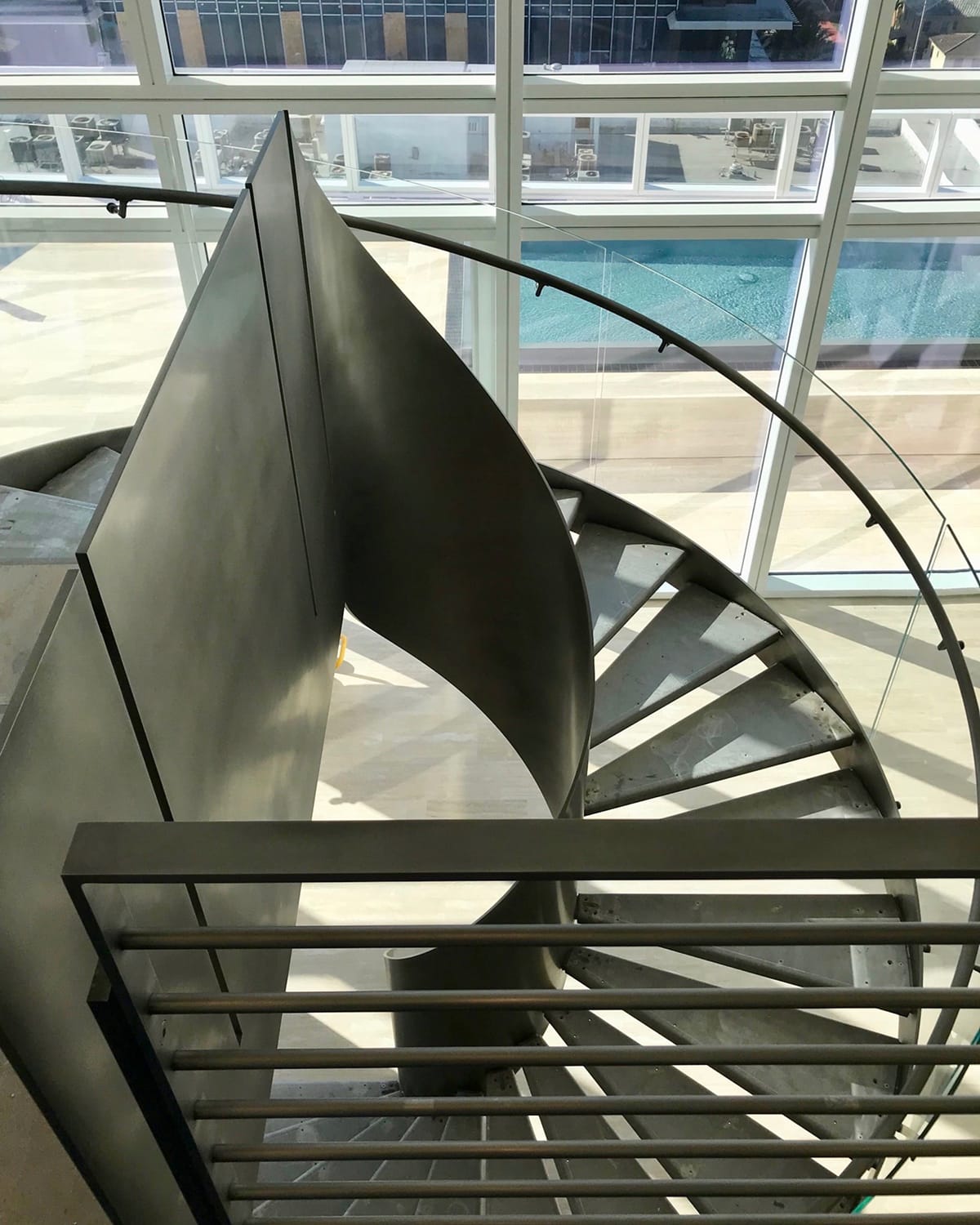 Spiral staircase with wood treads and a glass and brushed-metal railing, beside floor-to-ceiling windows