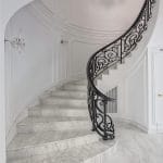 Spiral staircase with white marble treads and a scrolled iron railing, curving up from a marble landing in an all-white paneled rotunda