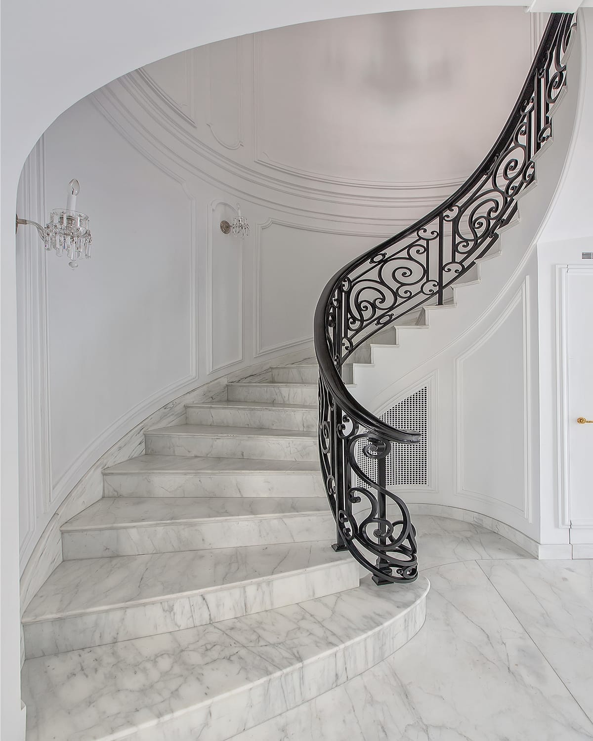 Spiral staircase with white marble treads and a scrolled iron railing, curving up from a marble landing in an all-white paneled rotunda