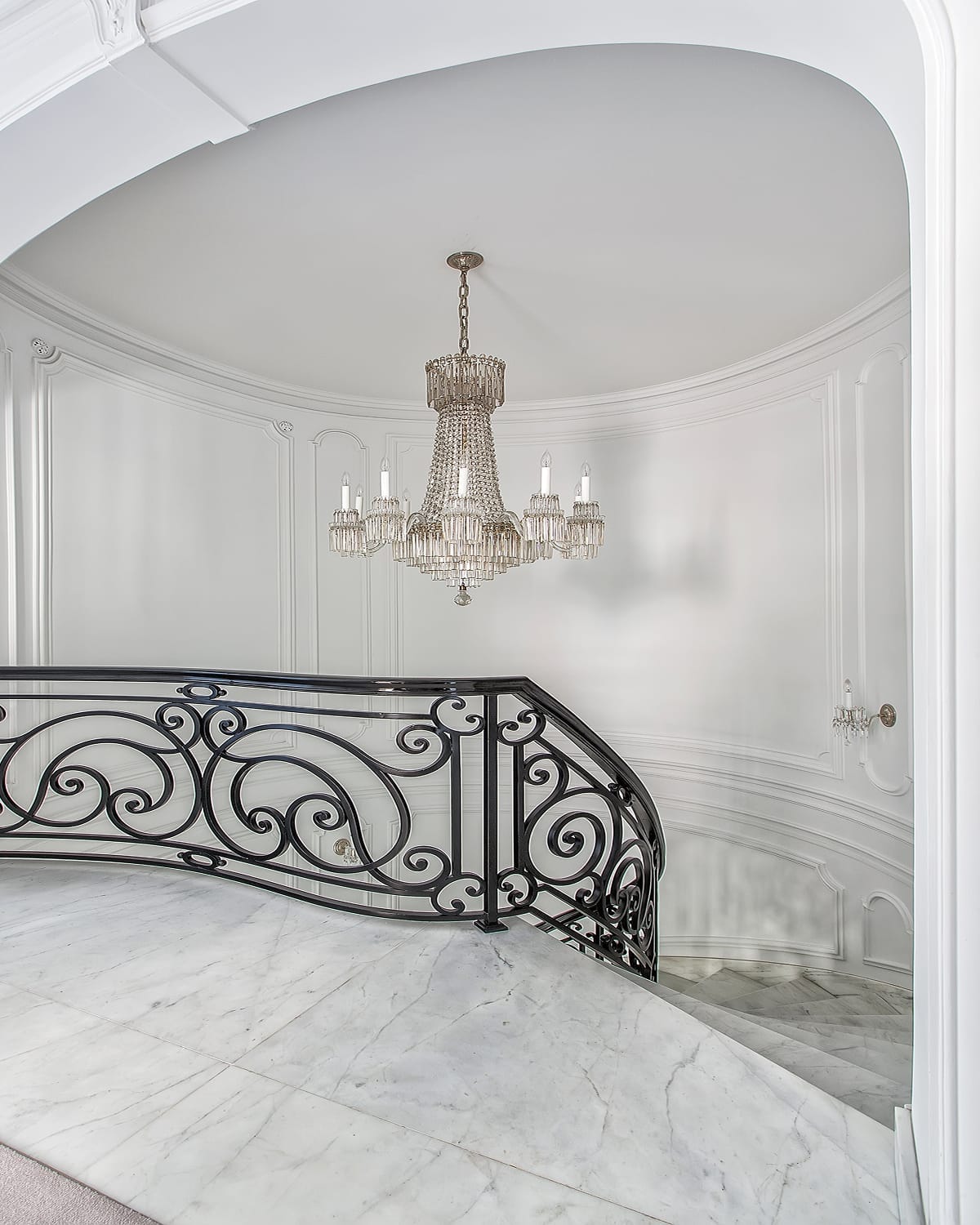 Top landing of a spiral staircase, with a scrolled iron railing framing the opening beneath a crystal chandelier and marble flooring