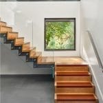 Floating staircase with wood treads edged in a rust-orange accent and a glass railing, next to a square window framing green foliage