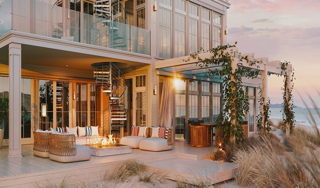 Coastal home exterior with a metal spiral staircase on the balcony, outdoor lounge seating, and a fire pit at sunset