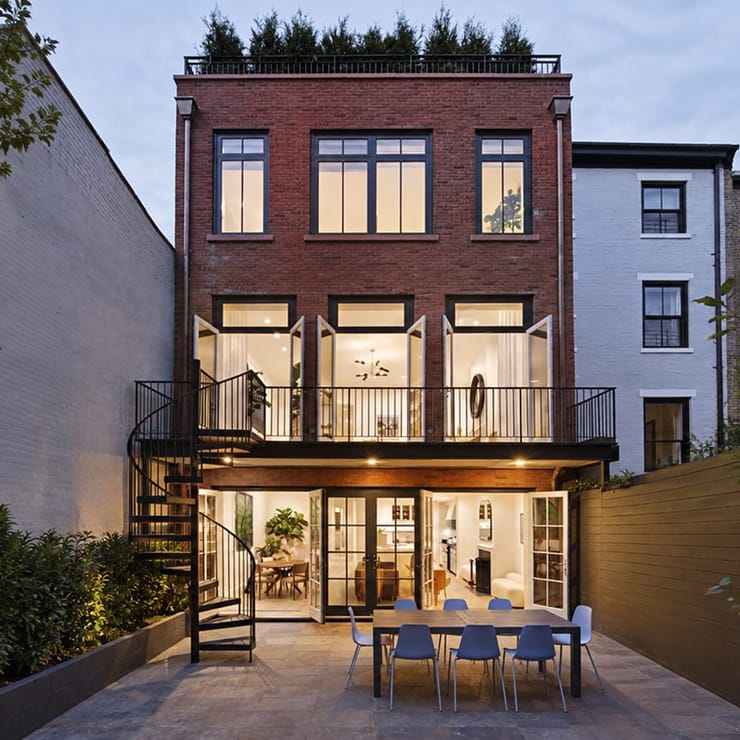 Black exterior metal spiral staircase on a brick rowhouse, connecting a balcony to a lit patio with dining table at dusk